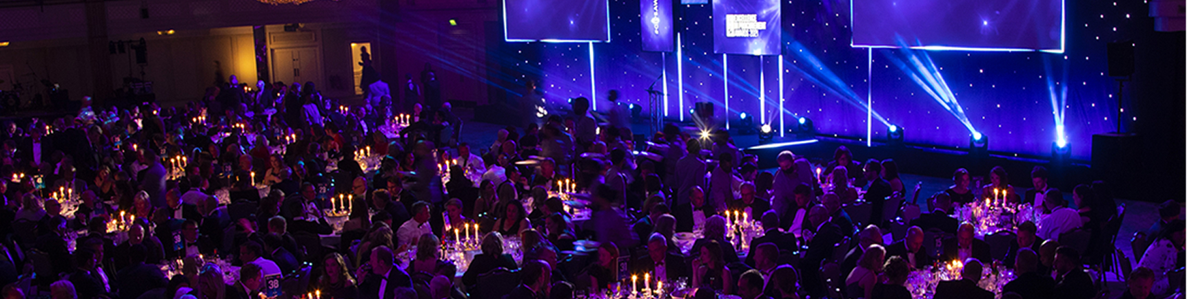 A wide, panoramic view of a large, dark, and elegant awards dinner. Many people are seated at candlelit tables in the foreground. In the background, a stage is dramatically lit with blue and purple spotlights and features large screens.