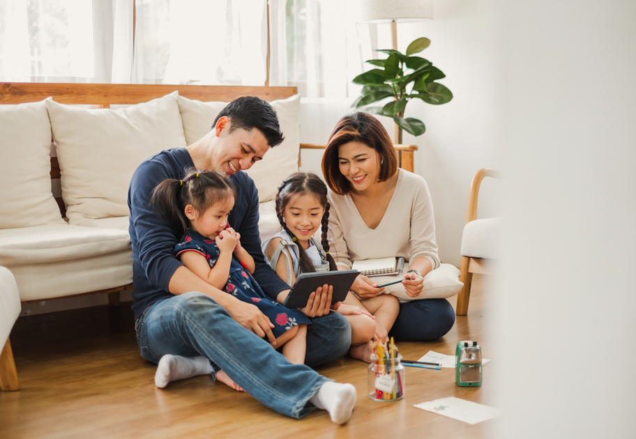 A family of four sits on the living room floor, smiling and looking at a tablet. Two young girls sit between their parents. Art supplies and paper are scattered nearby, and sunlight streams in from a window.