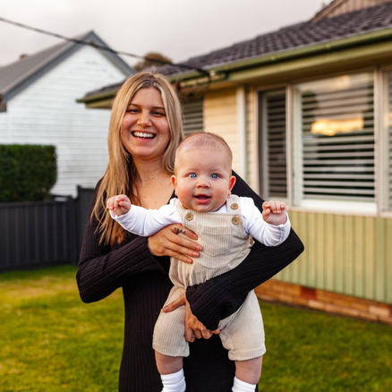 A smiling woman stands outside a house, holding a happy baby in light-colored overalls. The house and lawn are visible in the background on a sunny day.
