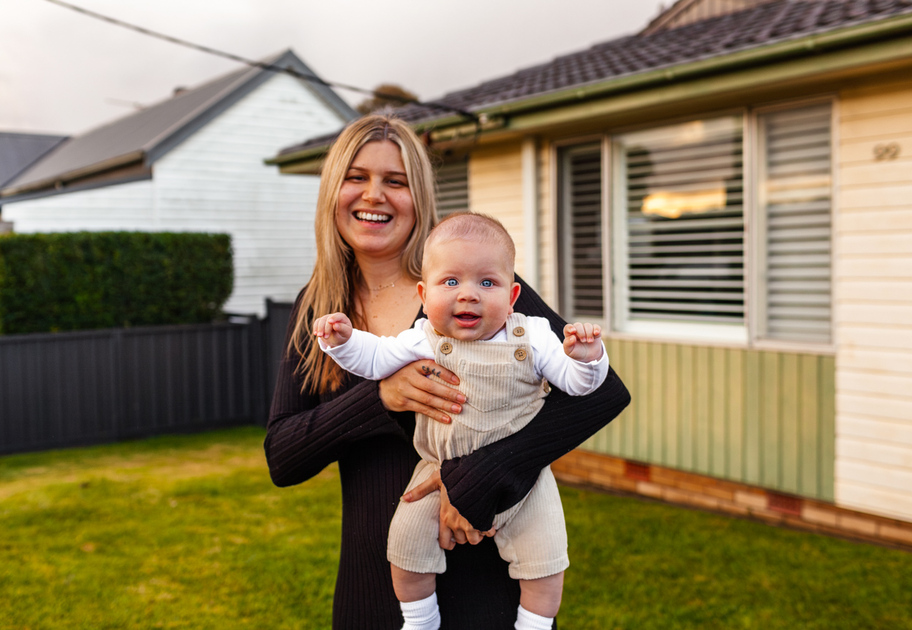 A smiling woman stands outside a house, holding a happy baby in light-colored overalls. The house and lawn are visible in the background on a sunny day.