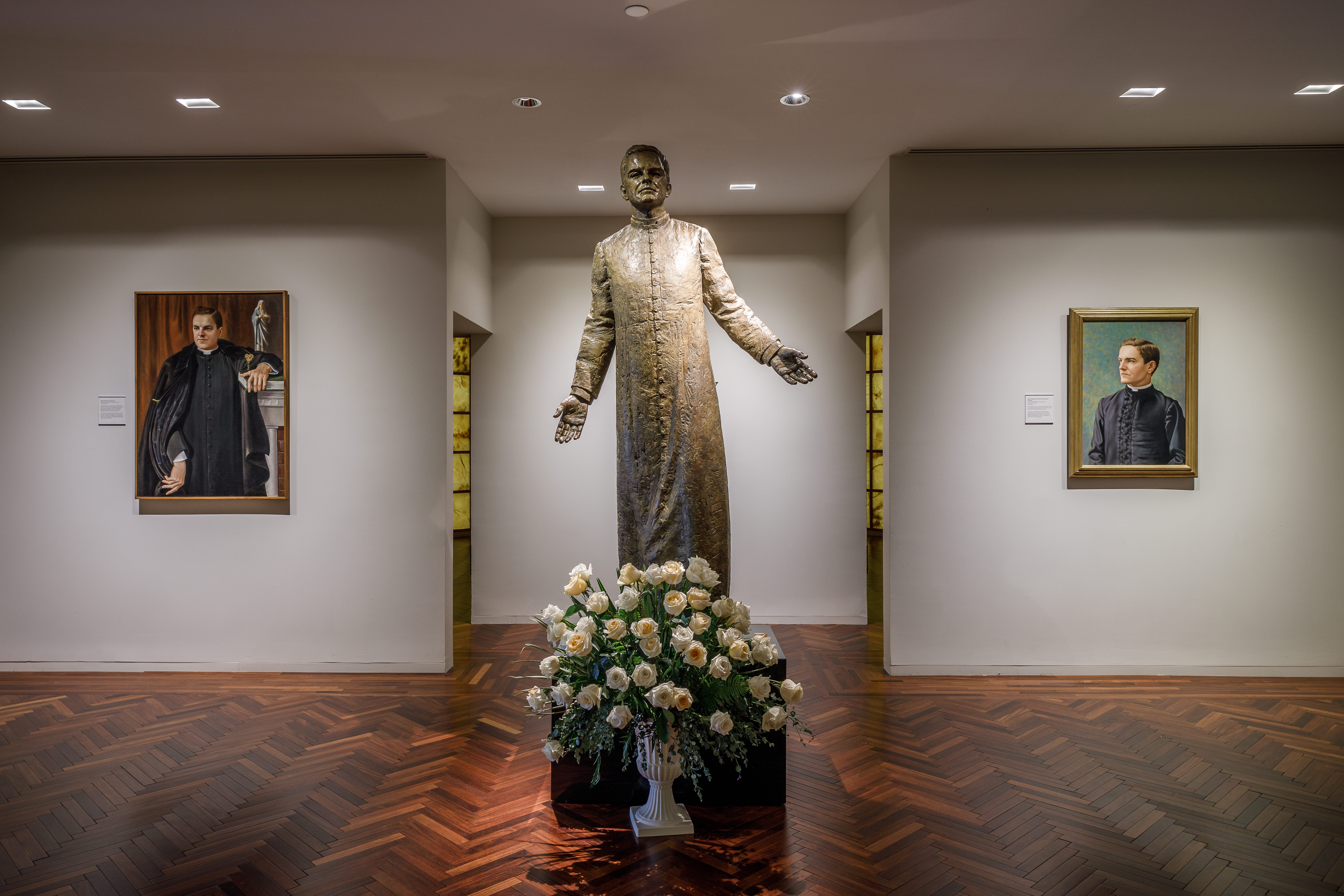 Statue of Father Michael J. McGivney in the McGivney Gallery at the Blessed Michael McGivney Pilgrimage Center, with portraits of him on either side.