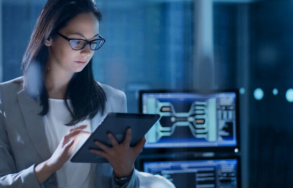 Woman standing in front of computers looking at a tablet