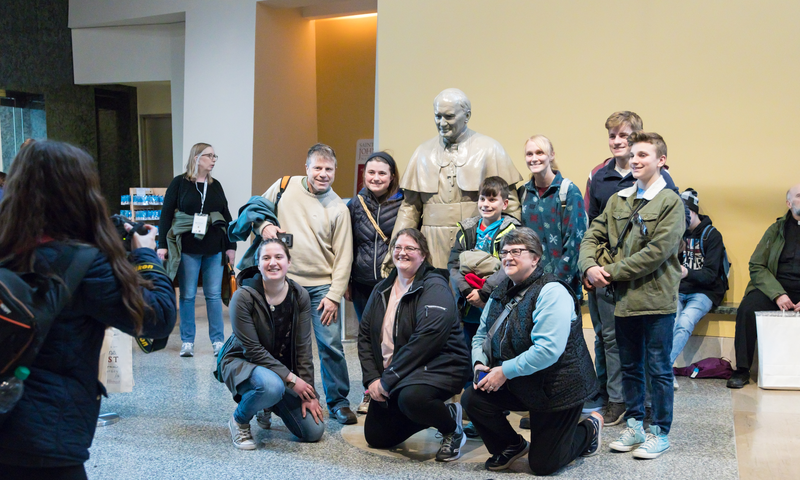 A group of people smiling and posing for a photo in front of a statue of JP2.