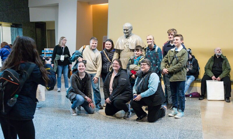 A group of people smiling and posing for a photo in front of a statue of JP2.