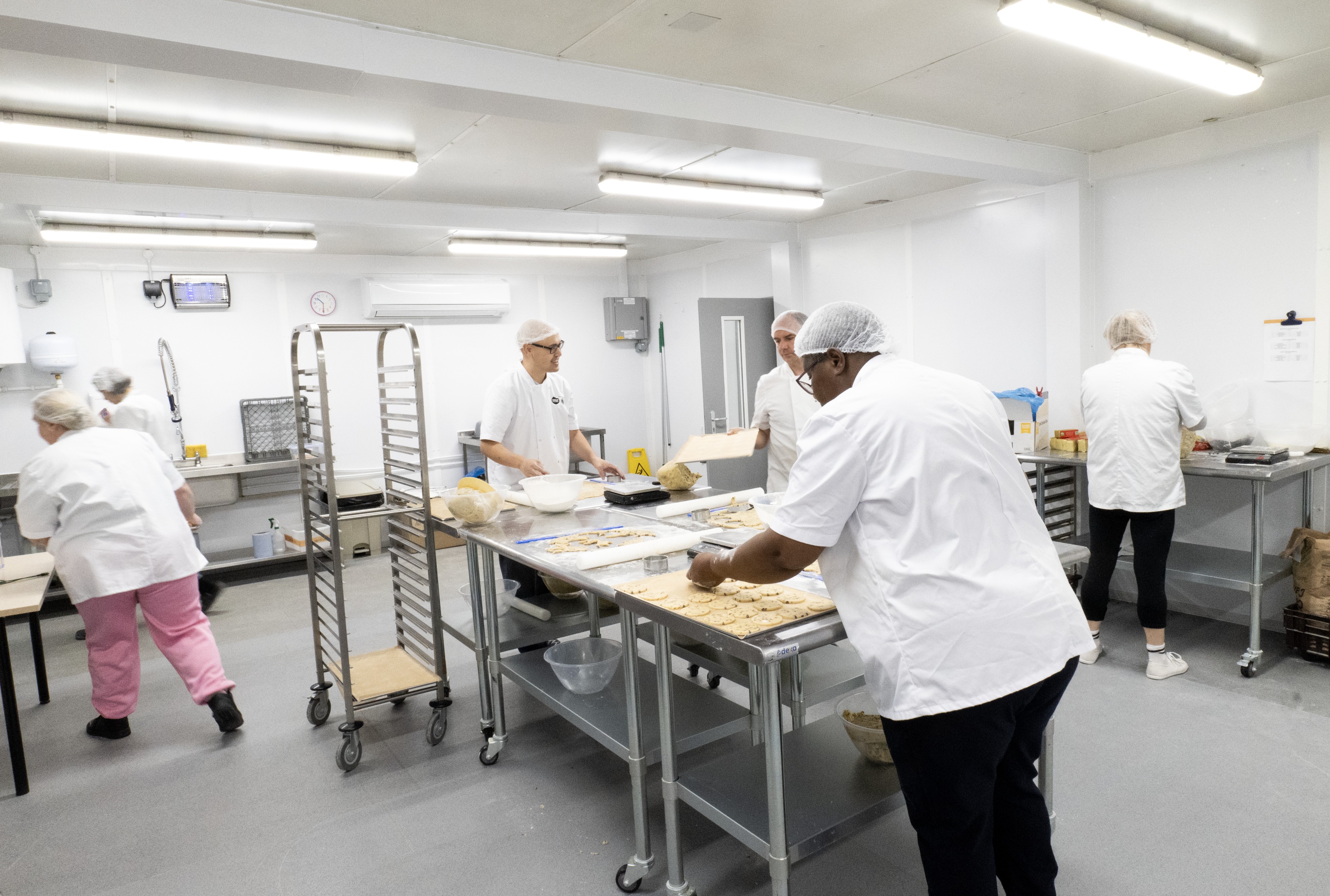Several bakers in white uniforms and hairnets work at stainless steel tables in a bright, modern commercial kitchen.
