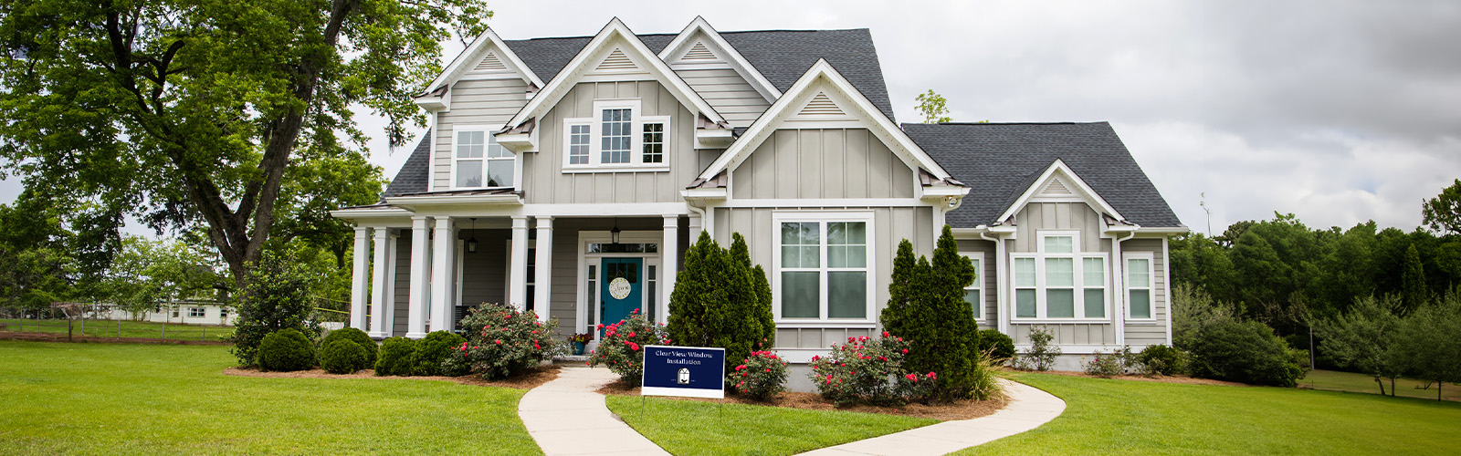 A nice two story house with grey siding sits on a well-manicured lawn with a large, beautiful tree beside it