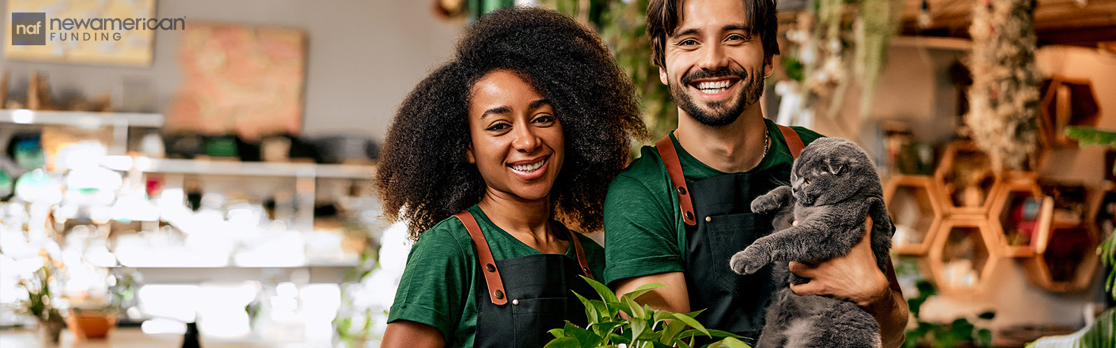A Black woman and a white man wearing aprons for work standing smiling together in a shop. The man is holding a cat. 