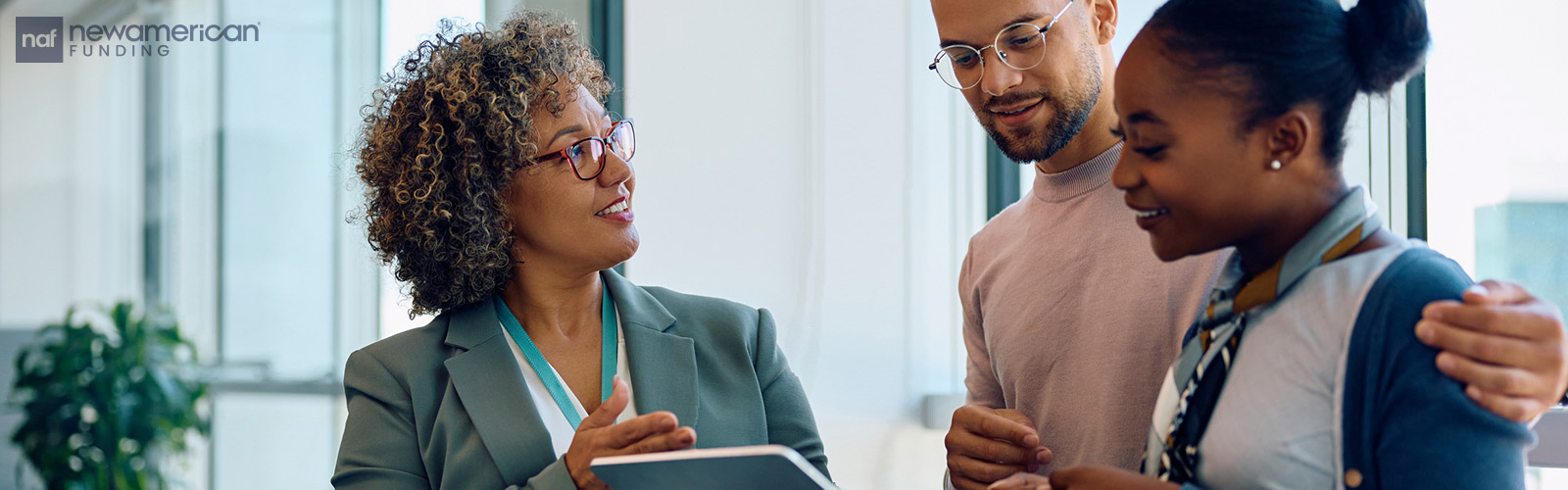 A Black woman dressed in a suite goes over something on a tablet with a smiling Black couple inside of a home