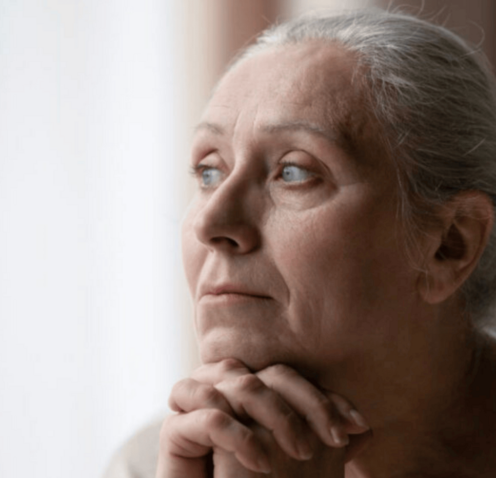 Older woman with grey hair sits by a window, resting her chin on her hands, looking thoughtfully outside with a calm expression.