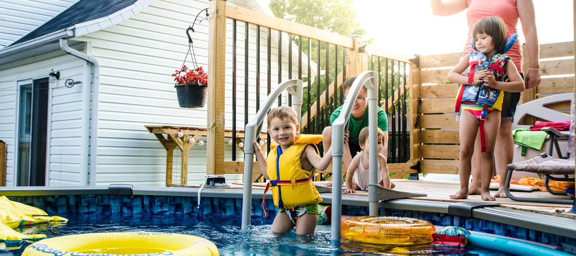 A little boy in a life jacket getting into a pool while his parents and sibling watch closely.