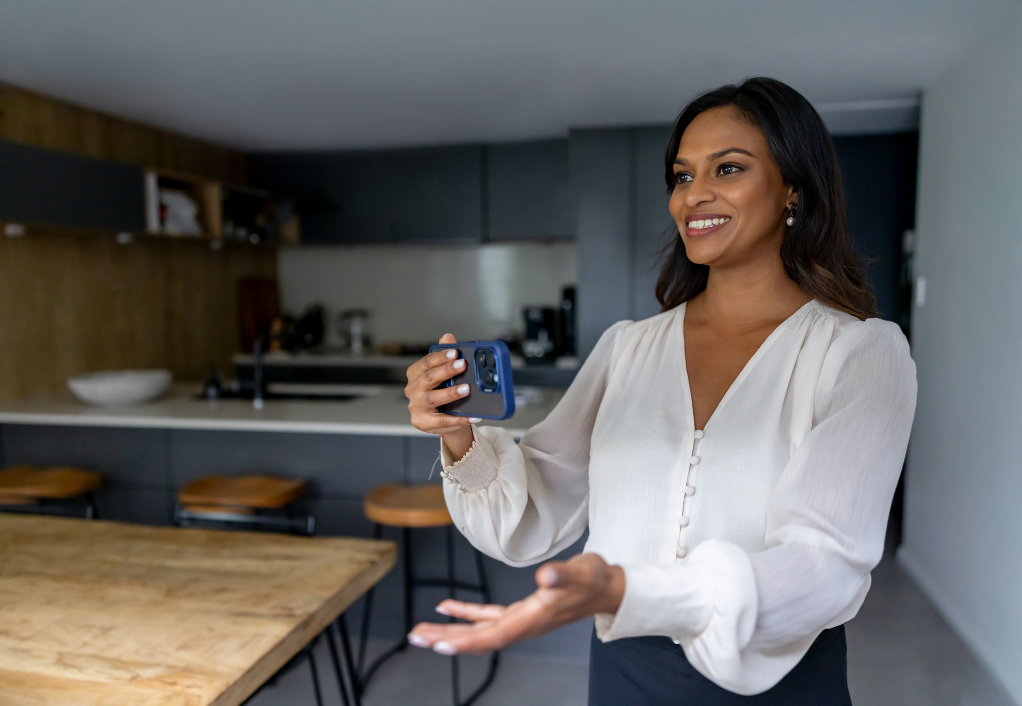 A smiling woman holding up her phone to film herself in a kitchen.