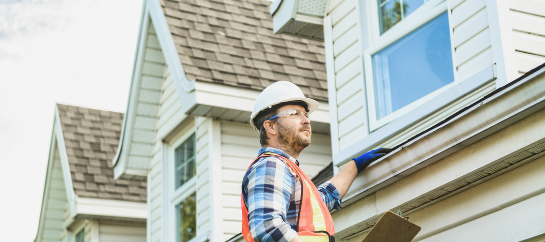 A home inspector on a ladder running a hand by the gutters on the roof of a home.