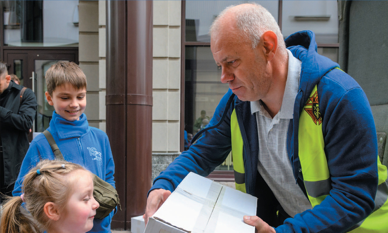 A Knight handing a girl a box with the Knights of Columbus logo on it, with a boy, smiling, standing in the background.
