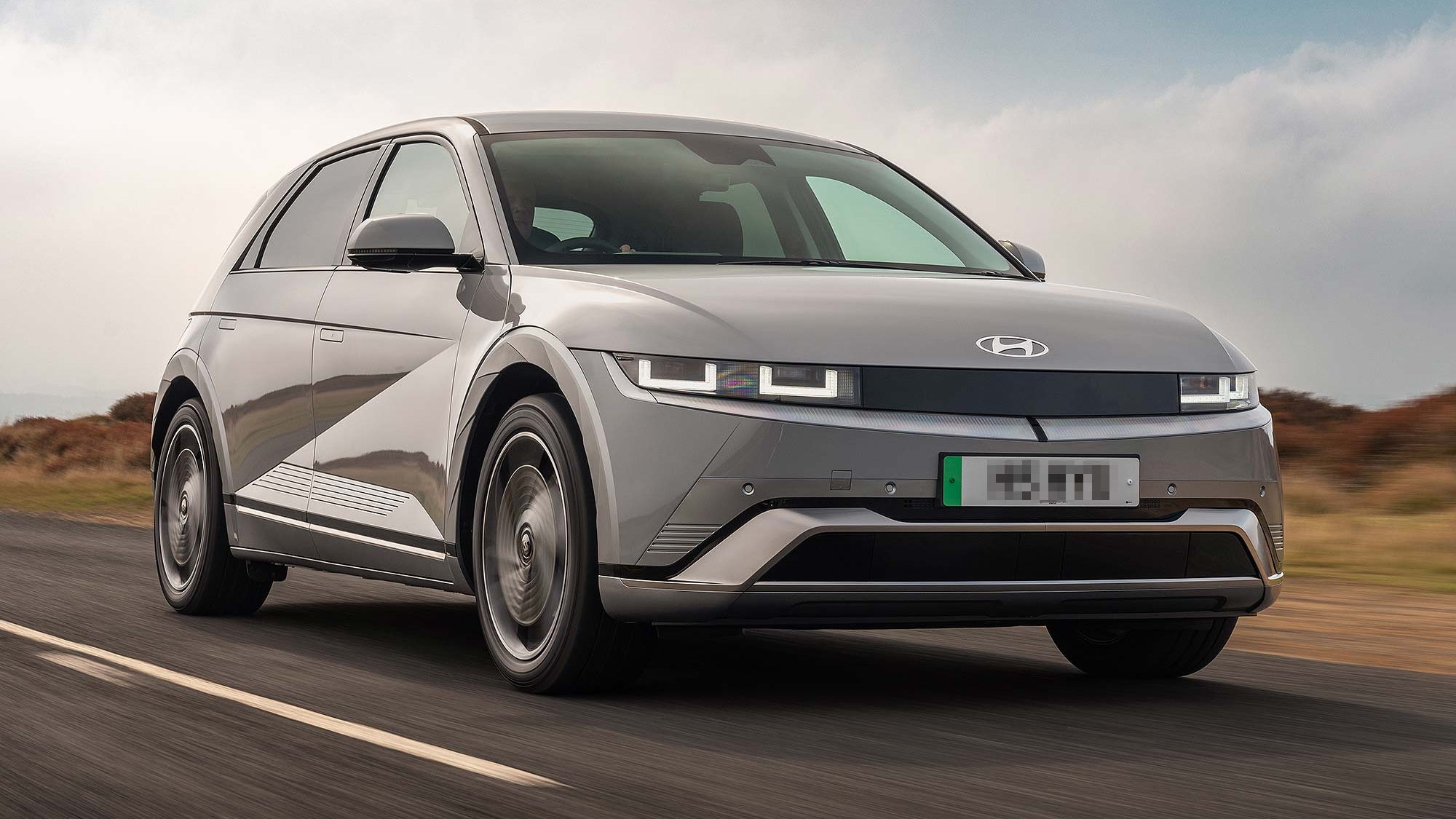 Grey Hyundai IONIQ 5 electric car driving on an open road, with countryside scenery and a cloudy sky behind.