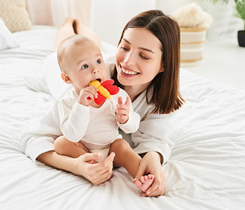 Mama na postelji z dojenčkom, ki izraščanje mlečnih zob lajša z grizalom