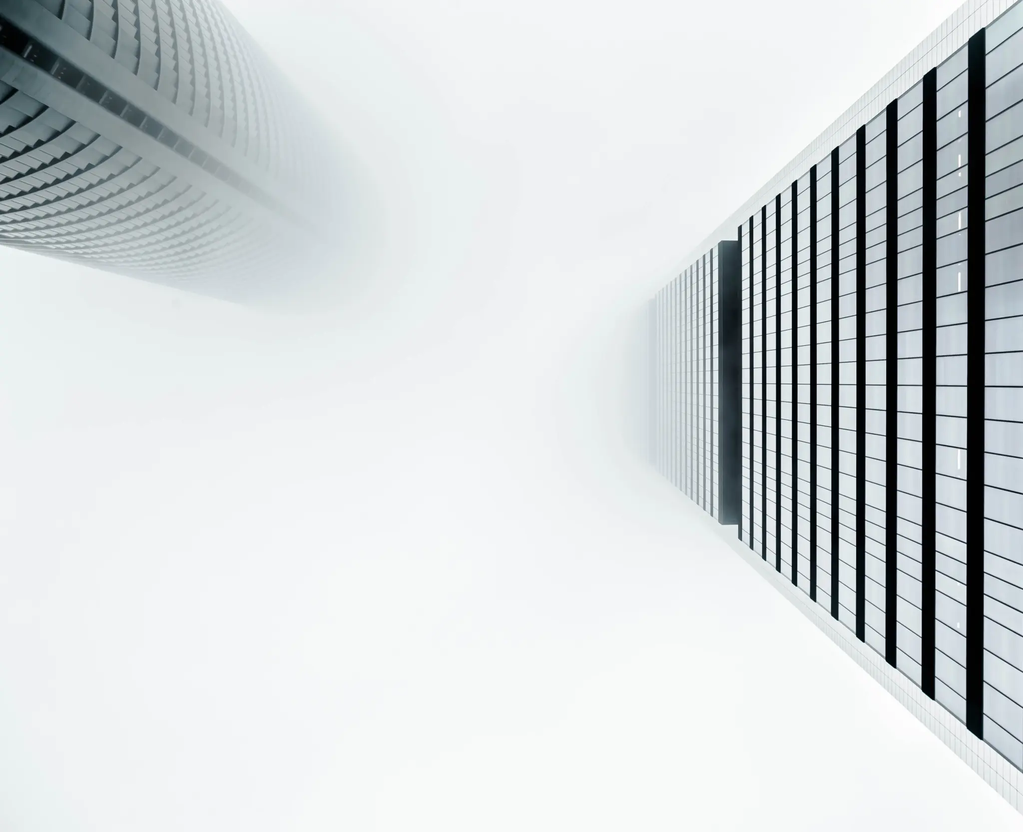 A minimalistic upward view of two modern skyscrapers disappearing into thick white fog. The building on the left is cylindrical with horizontal window patterns, while the one on the right is rectangular with vertical and horizontal lines forming a grid pattern. The fog obscures the tops of both buildings, creating a surreal and abstract atmosphere.