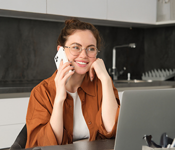 Mlada ženska v kuhinji sedi za laptopom in govori po telefonu - simbolni prikaz fleksibilnega dela