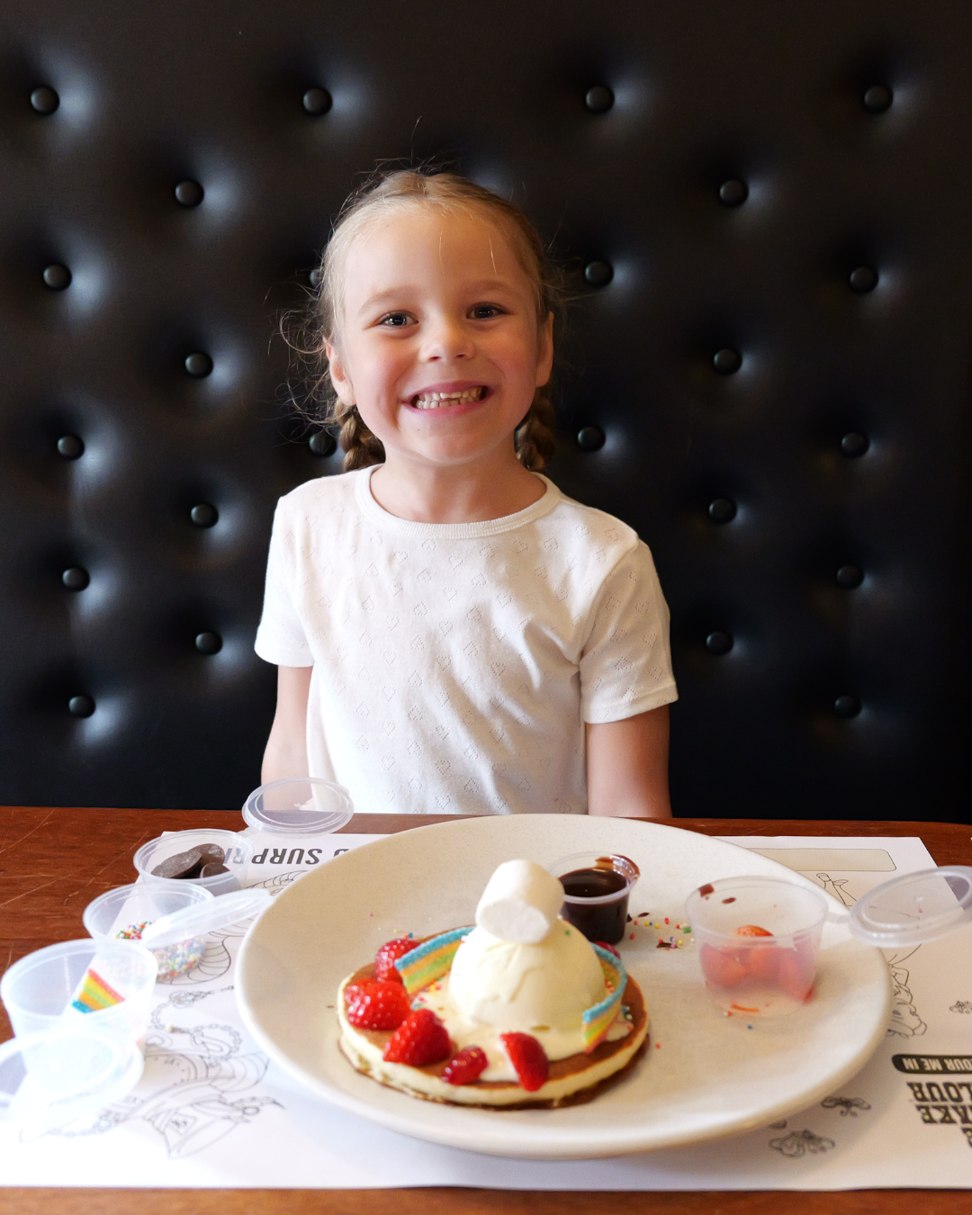A child sitting in a restaurant booth decorating a pancake topped with ice cream, strawberries, marshmallows and colourful toppings, with small containers of sprinkles and sauce on the table.