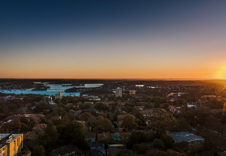 Aerial view of a suburban area with houses, trees, and buildings at sunset. The sun is low on the horizon, casting an orange and blue gradient across the sky and reflecting off water in the distance.