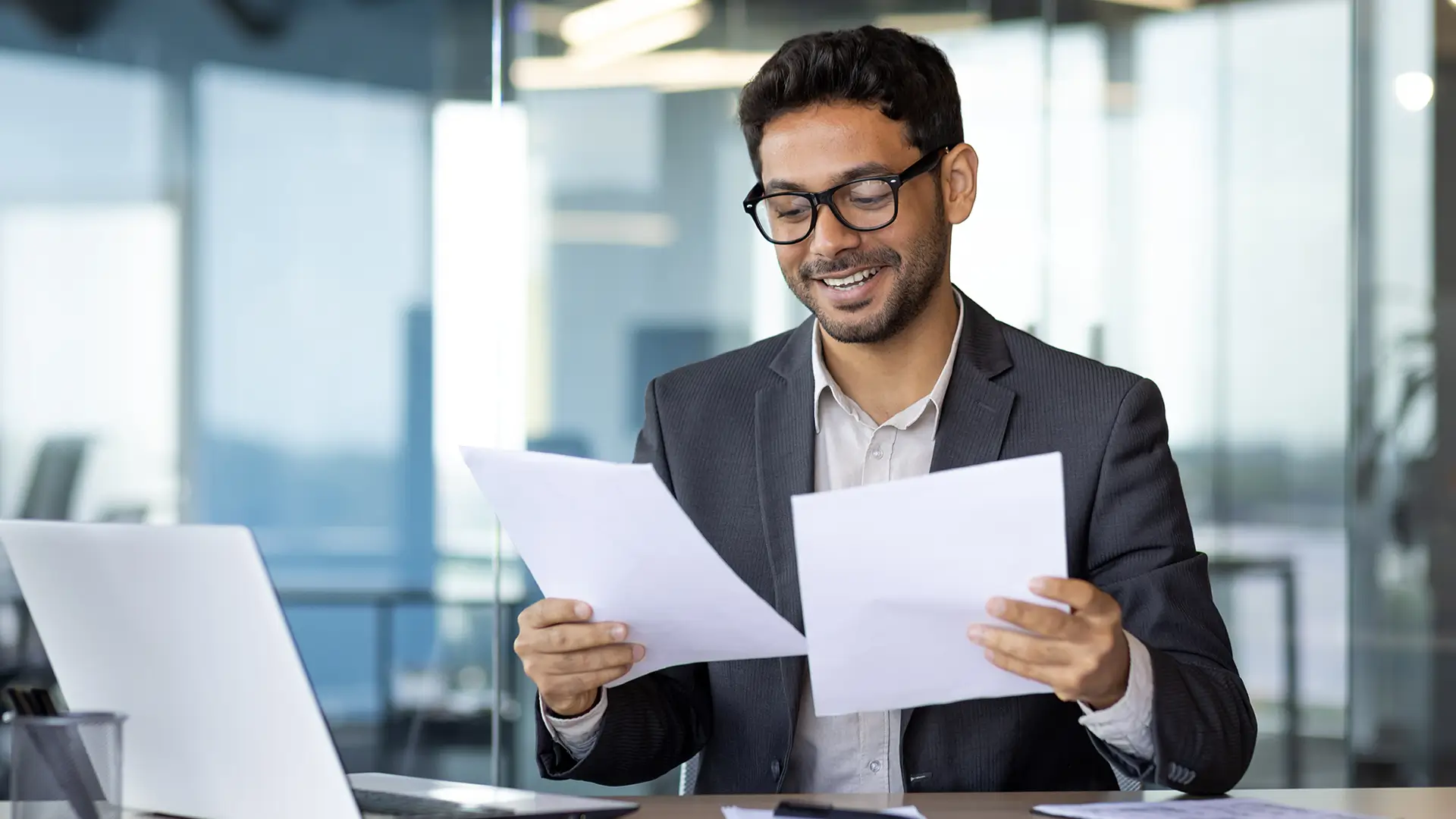 Smiling professional reviewing documents at an office desk with a laptop.
