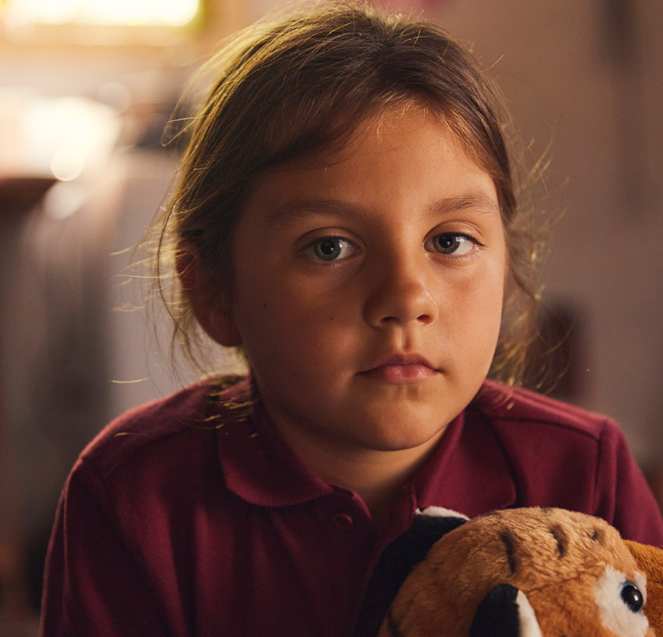 A young girl with brown hair and a maroon shirt looks at the camera with a serious expression, holding a plush toy in a softly lit indoor setting.