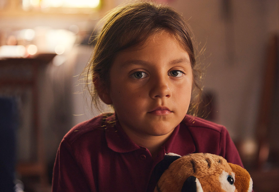 A young girl with brown hair and a maroon shirt looks at the camera with a serious expression, holding a plush toy in a softly lit indoor setting.