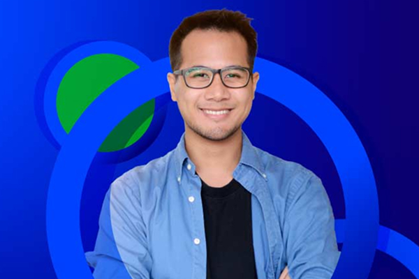 A young man wearing glasses and a blue shirt over a black t-shirt smiles at the camera, set against a background of concentric blue and green circles.