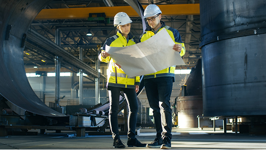 
Two workers, a man and a woman, wearing hard hats and high-visibility jackets, are standing inside a large industrial factory or heavy manufacturing facility. They are looking intently at a large blueprint or technical drawing they are holding between them. Large metal structures and heavy machinery are visible in the background.