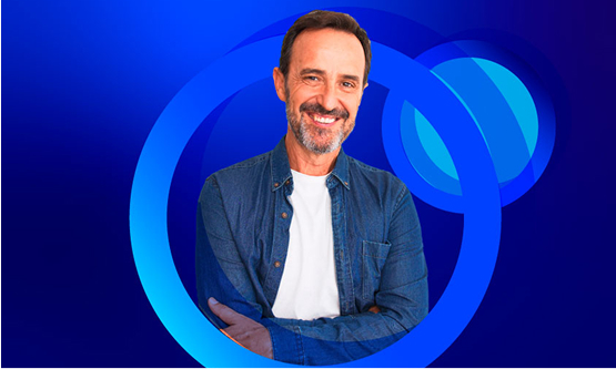 A smiling man with a beard and a denim shirt over a white t-shirt stands with his arms crossed. The background is a bright blue with concentric blue circles.