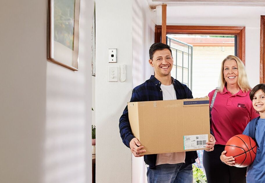 Mission Australia staff member supports a smiling family arriving at their new home with moving boxes and a basketball.