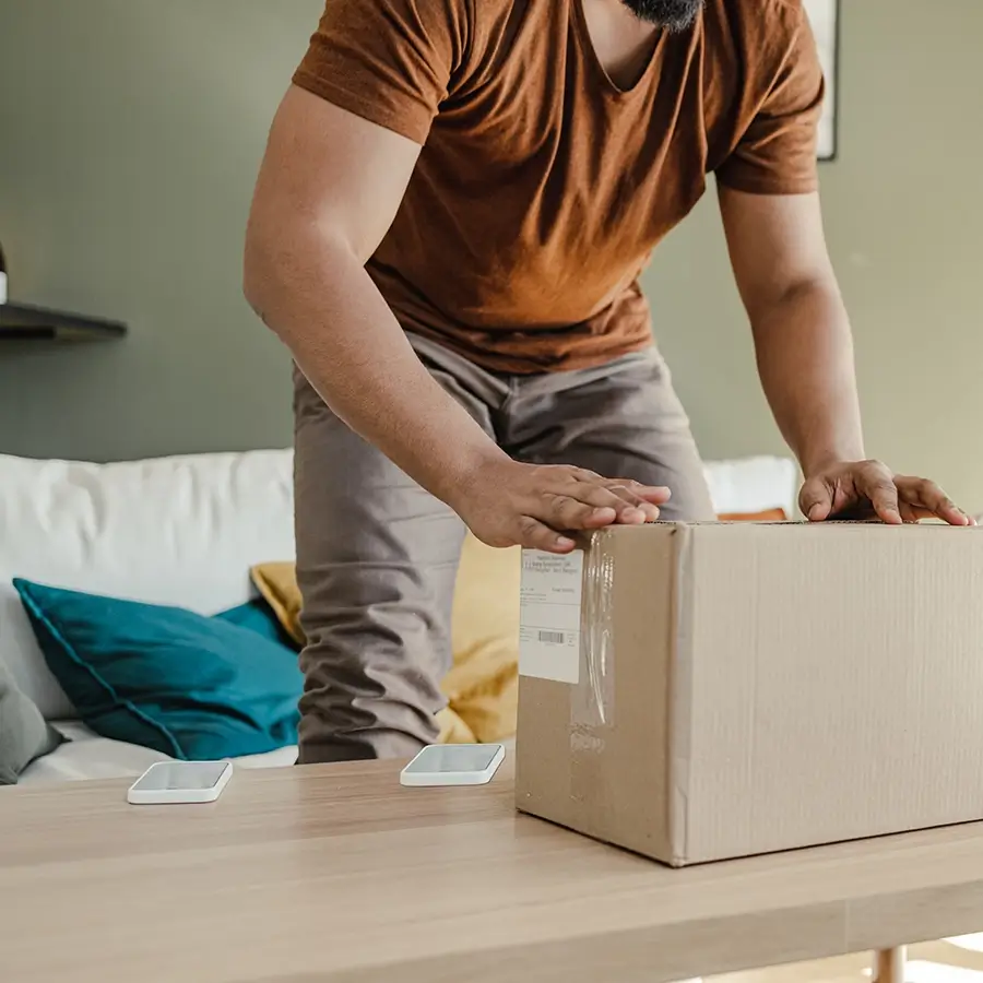 modern-om-cost-optimizer-bus-outcomes-900x900.webp Man placing or retrieving a cardboard shipping box from a wooden coffee table in a living room, with colorful pillows and smartphones nearby, illustrating home delivery.