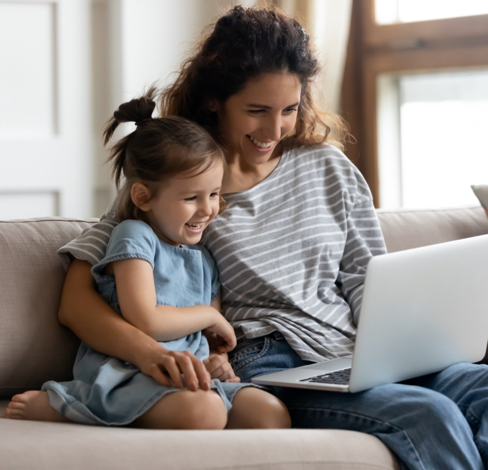 	
A smiling woman and a young girl sit together on a couch, looking at a laptop and laughing. They both wear casual clothes and appear to be enjoying their time together in a bright, cozy living room.