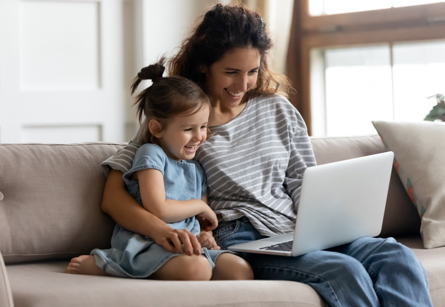 A smiling woman and a young girl sit together on a couch, looking at a laptop and laughing. They both wear casual clothes and appear to be enjoying their time together in a bright, cozy living room.