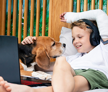 Deček s psom leži v postelji in gleda serije na računalniku. Roko ima v mavcu, ki je posledica padca - ene najpogostejših nevarnosti za otroke.