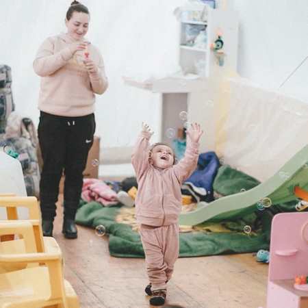 A young Ukrainian girl and her mother play with bubbles in a playroom in a refugee camp during war times.