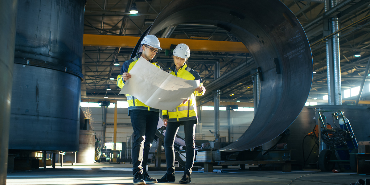 Two workers in hi-vis jackets and hard helmets looking at a plan together, standing in a factory environment