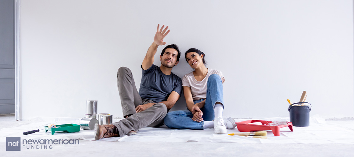 A couple sitting on a drop cloth with paint supplies around them in an empty room.