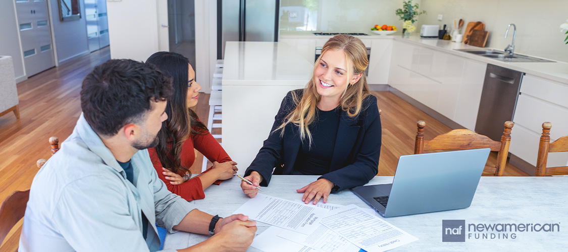 A couple meeting with a smiling real estate agent at a table with paperwork in front of them.