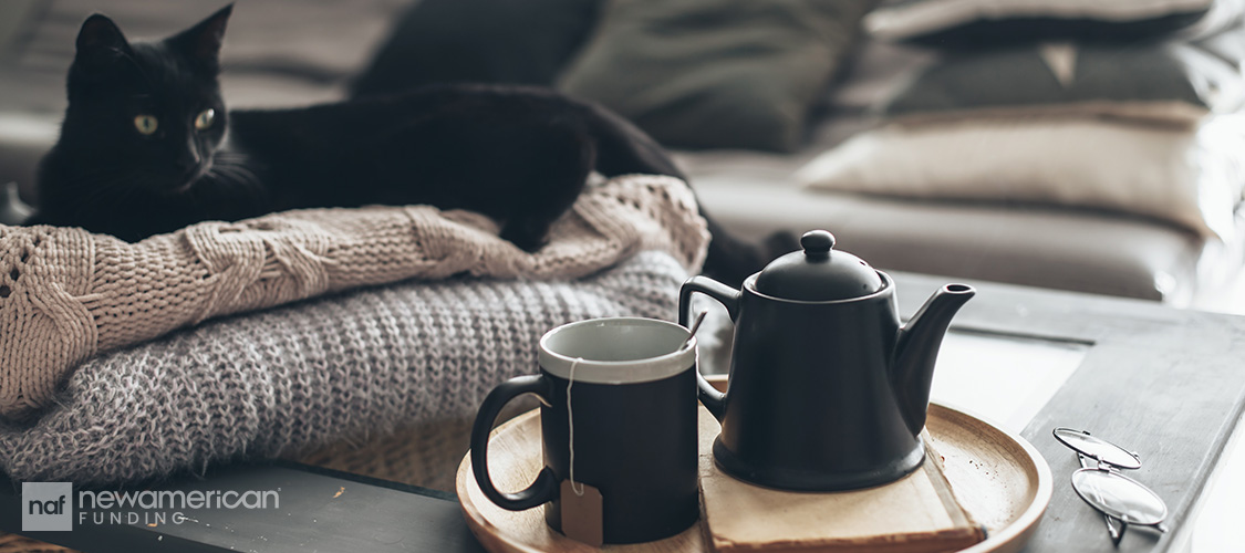 A black cat lying on blankets next to a tray with a tea kettle and a mug.