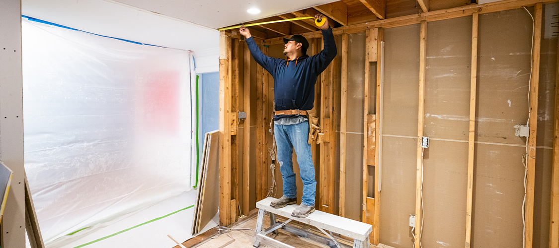 A man measuring a wall that hasn't been finished in a building.