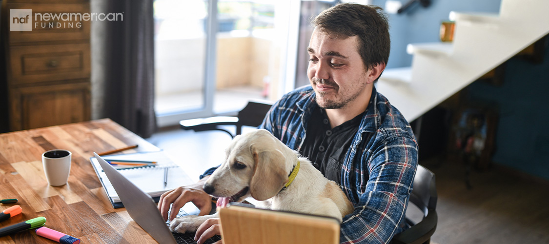 A man sitting at a table with a dog on his lap looking at a laptop.