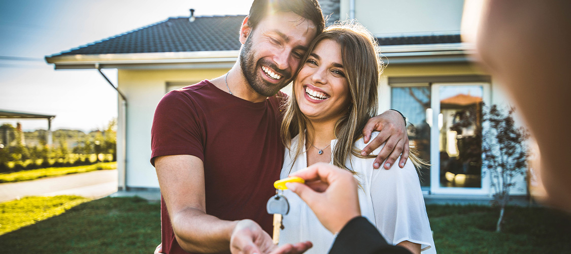 A couple standing outside of a house with someone reaching out to give them the keys.