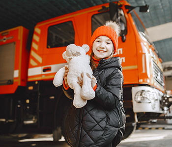 Deklica s plišastim medvedkom na obisku gasilske postaje spoznava delo gasilcev