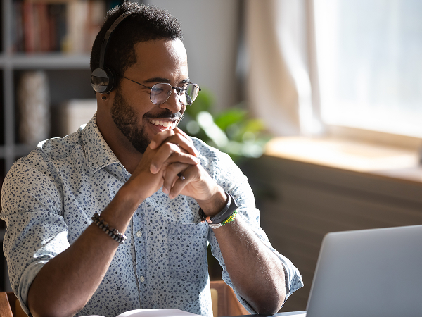  A smiling man wearing headphones and glasses looks down at a laptop screen with his hands clasped in front of him. He is wearing a light blue patterned shirt.