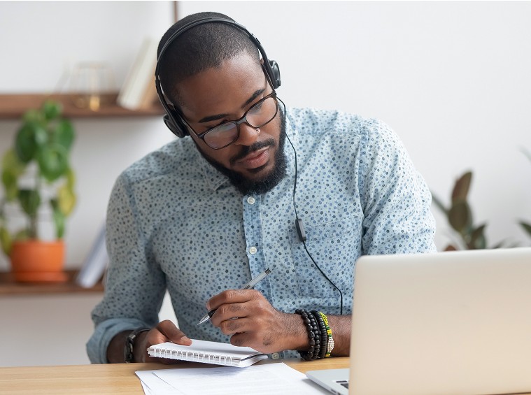 Man wearing headphones studying online and taking notes in a notebook