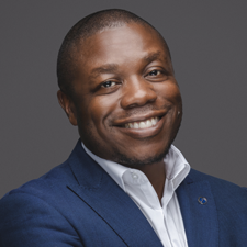 Portrait of Sam Achampong smiling with arms crossed, wearing a blue blazer over a white collared shirt against a dark grey background. profile image