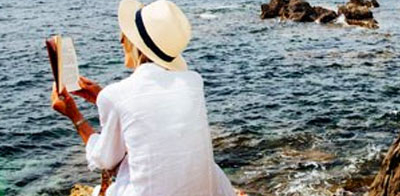 A woman sits on a rock by the ocean, reading a book, representing a female solo travel guide blog.