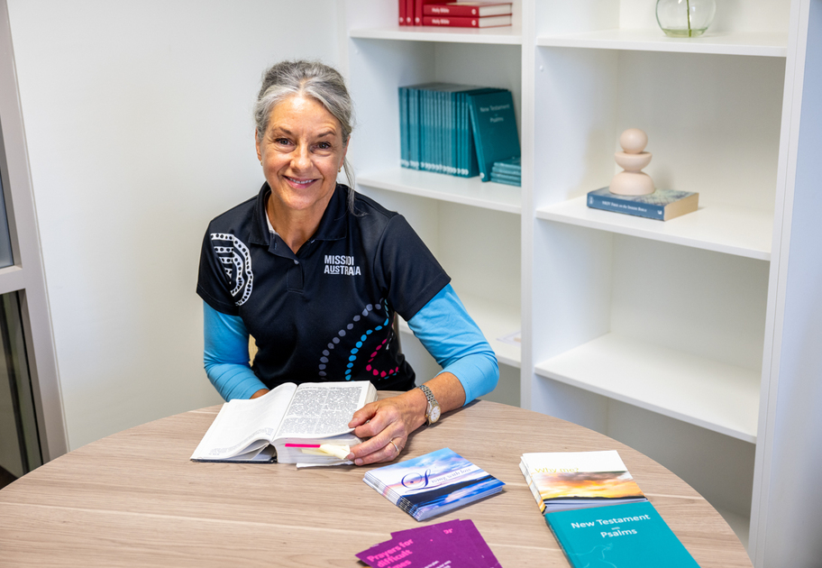 A woman with gray hair, wearing a Mission Australia shirt, sits at a round table reading an open book. Various books and pamphlets are spread on the table. Shelves with books and decor are in the background.