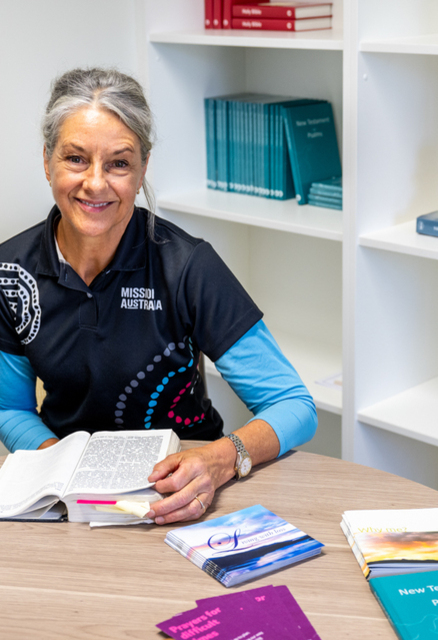 A woman with gray hair, wearing a Mission Australia shirt, sits at a round table reading an open book. Various books and pamphlets are spread on the table. Shelves with books and decor are in the background.