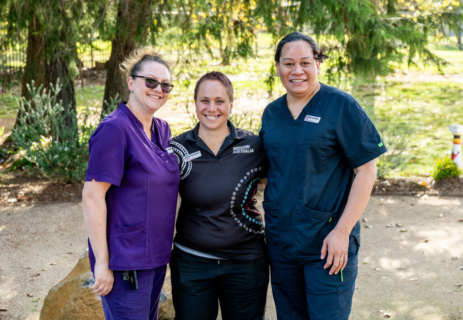 Three women stand outdoors smiling, dressed in Mission Australia uniforms—two in scrubs (purple and navy blue) and one in a black polo shirt. Green trees and sunlight fill the background.